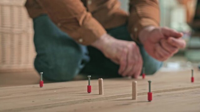 Skilled individual carefully positions wooden pegs and screws on a wooden floor, demonstrating precise assembly techniques in a well-lit indoor workspace with tools nearby