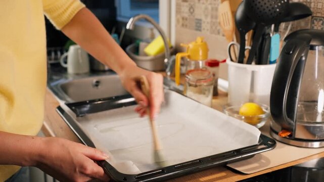 Close up of woman hands use pastry brush to oil parchment on baking sheet, greasing for homemade baking in warm, cozy home kitchen during meal preparation