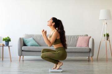 Obraz premium Full length of excited Indian woman sitting on scales at home, overjoyed with success of her slimming diet, side view. Emotional Asian lady achieving her weight loss goal, making YES gesture