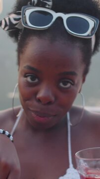 Vertical portrait shot of African American woman, holding glass of wine, smiling joyfully at camera as she dancing during lakeside celebration with her friends