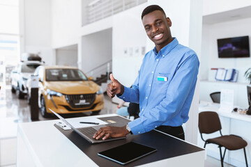 Fototapeta premium Portrait of smiling black car salesman showing thumb up gesture while using laptop at automobile dealership. Cheerful Afro manager posing at workplace, looking at camera in auto showroom