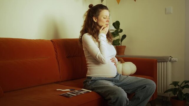 Unwell pregnant woman sitting on the sofa, suffering from morning sickness and toxicosis as she clutches her belly and chest, with ultrasound photos and a pregnancy test lying next to her