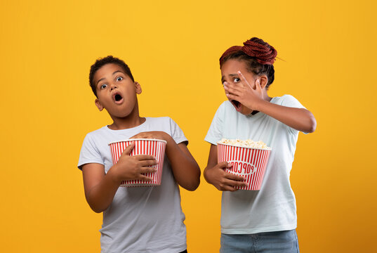 Frightened afro-american boy and girl siblings or friends with popcorn buskets watching horror, sister closing her face, bother grimacing, children watching spooky movie together, yellow background
