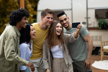 Obraz premium Positive multiracial friends taking selfie near their trailer, smiling and having fun together during autumn camping trip. Cheerful young people making photo of themselves at campsite