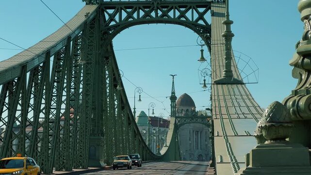 Scenic view of the green Liberty Bridge in Budapest, Hungary, over the Danube. Traffic with yellow trams crosses the historic steel structure under a clear sunny blue sky.

