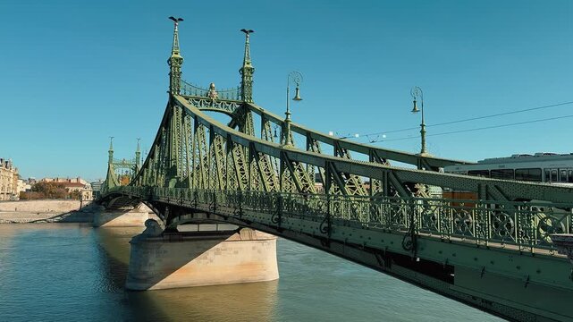 Scenic view of the green Liberty Bridge in Budapest, Hungary, over the Danube. Traffic with yellow trams crosses the historic steel structure under a clear sunny blue sky.

