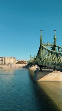 Scenic view of the green Liberty Bridge in Budapest, Hungary, over the Danube. Traffic with yellow trams crosses the historic steel structure under a clear sunny blue sky.

