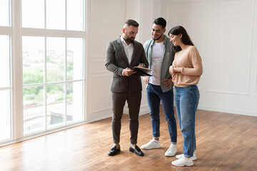 Smiling young married couple discussing agreement with skilled real estate agent or broker. Professional financial advisor or salesman explaining contract details with interested clients at meeting