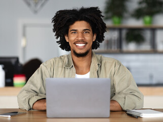 Cheerful young handsome african american curly male working at laptop at table with notepad and smartphone in cafe interior. Study and work with modern technology and modern digital nomad at workplace