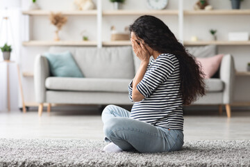 Pregnancy hormonal changes concept. Young curly long-haired pregnant woman feeling down, sitting on floor in cozy living room and crying, staying alone at home, side view, free space © Prostock-studio