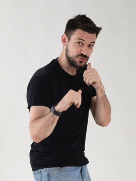 Expressive young man in black t-shirt standing in a defensive boxing pose, looking at camera with funny defiant face, grey background.