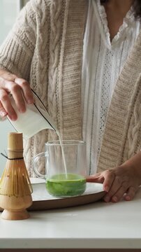 Woman pouring milk into a glass with matcha green tea for a healthy drink. Preparing matcha green tea latte at home.