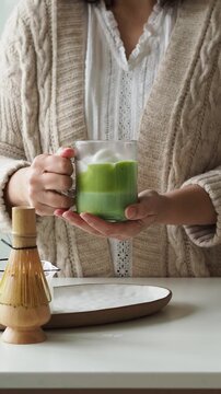 Female hands holding a glass of fresh matcha latte with foam on a white table