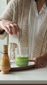 Woman adding foamed milk to a glass cup with matcha green tea to prepare a healthy drink.