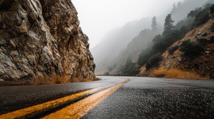Winding wet mountain road with yellow lines leading through foggy rocky landscape.