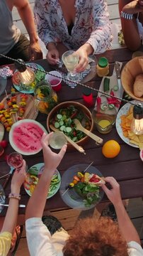 Vertical top view shot of multiethnic group of young friends, sitting at dinner table on wooden pier for lake party, cheerfully clinking their glasses in toast