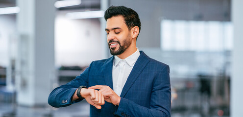 A man in a suit looks at his watch while standing in a modern office. The setting shows a workspace with bright lighting and a clean design. He appears focused and engaged in his task.