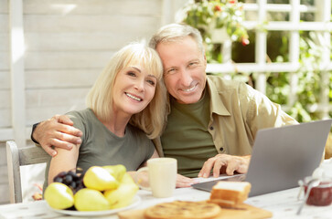 Happy senior couple using laptop computer outdoors, sitting at home terrace and smiling to camera. Retired man and woman using modern device. Elderly family reading news, shopping online