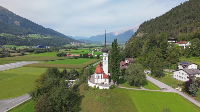 A small church with a pointed bell tower in a valley in Austria. Aerial view of the religious site against the backdrop of the mountains and landscape of the Oberes Inntal valley in Tyrol
