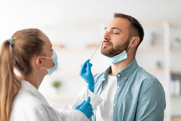 Female doctor making nasal coronavirus PCR test for millennial guy at clinic. Young Caucasian man making covid diagnostic procedure. Infectious disease protection concept