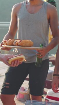 Vertical tilt-up shot of group of smiling male friends grilling hot dogs and BBQ sausages at lakeside gathering