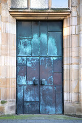 Weathered metal double door set in historic stone building facade with tall window above, aged surface texture showing time passage, closed entrance expressing mystery, strength