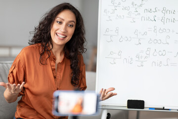 Smiling female sitting in front of smartphone on stabilizer, recording video or having live videoconference with students. Happy young smart woman filming educational lecture, explaining theme