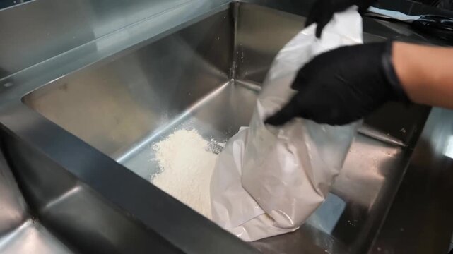 A kitchen worker wearing black gloves uses scissors to open a white bag and pours fine flour into a professional stainless steel container before mixing it manually for food preparation