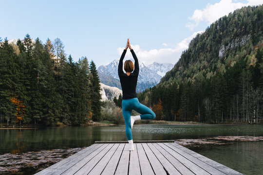 Mature fit woman practicing yoga on pier by lake overlooking green nature and mountains. Beautiful female in harmony with nature outdoors. Concept of wellbeing, life balance, mindfulness, zen energy.