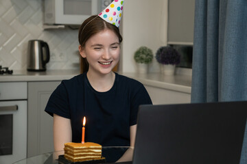 Happy teenager wearing party hat celebrating birthday online using laptop, sitting at kitchen table with small piece of cake and candle, making video call with family or friends
