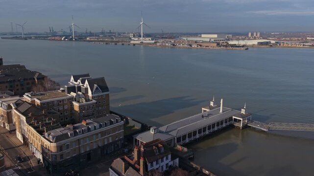 Above historical Gravesend Pier on river Thames. Aerial view of Gravesend town and the pier with spinning wind turbines in the background at Tilbury Docks. Two videos in one with side and front view.