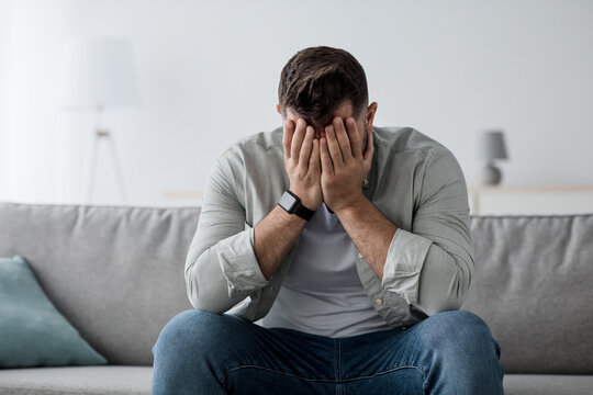 Frustration, mental disorder, broken heart. Mental problems, middle age crisis. Sad upset handsome bearded male sitting on couch crying, covers face with hands in living room interior, copy space