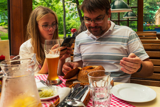 Father and young daughter sit at a restaurant table with bread, drinks, and tableware, looking at a smartphone while ordering or checking directions. Family dining concept, modern travel routine, and 