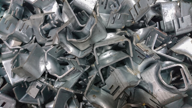 Metal industrial brackets stacked in bulk showing raw texture, manufacturing detail, silver surface, repetition, pattern, and metallic tones.