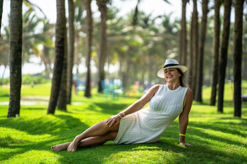 A girl in a white dress and hat lies on a green lawn against a background of palm