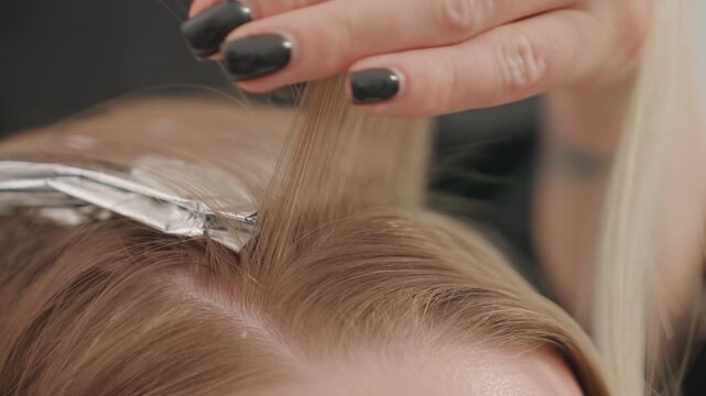 Closeup caucasian woman hair highlighting with foil in salon, stylist sections fine blonde strands, applies lightener with brush, folds foil over roots, smooths strands near scalp, neutral studio