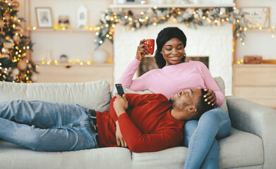 Obraz premium A couple sits on a couch during the holiday season. The woman smiles while holding a cup, and the man looks relaxed as he rests his head in her lap.
