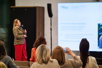 Obraz premium Audience watches presenter at conference. Woman on stage speaks into microphone. Attendees listen and take notes. One person records with smartphone.