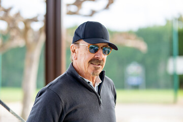 A man wearing sunglasses and a black hat stands in front of a tree
