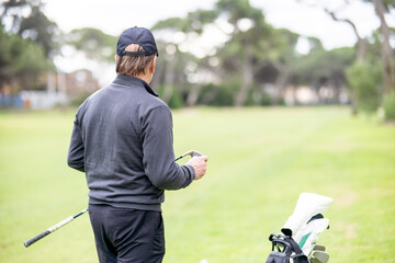 A man in a grey jacket is holding a golf club and looking at his phone