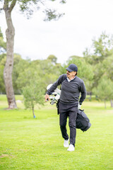 A man in a black jacket and hat is walking on a grassy field with a golf bag