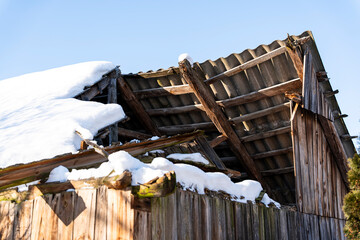 Damaged wooden barn roof partially covered with snow against a clear blue sky, emphasizing the contrast between winter elements and structural decay. Ideal for themes of abandonment or rural life. © daily_creativity