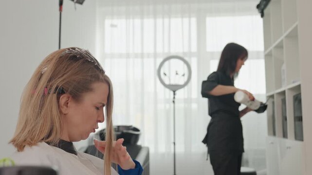 woman in salon chair getting highlights, stylist preparing products and checking cabinet, ring light and window backlight, foils and bleach application, client examining sectioned strand while stylist