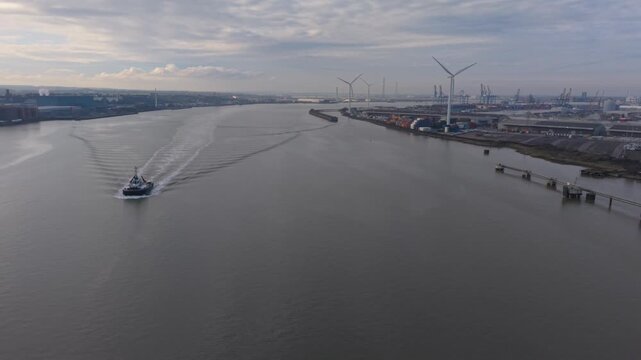 Pilot boat sailing on the river Thames aerial view. Spinning wind turbines at Tilbury Docks in the background. Industrial skyline on a cloudy day.