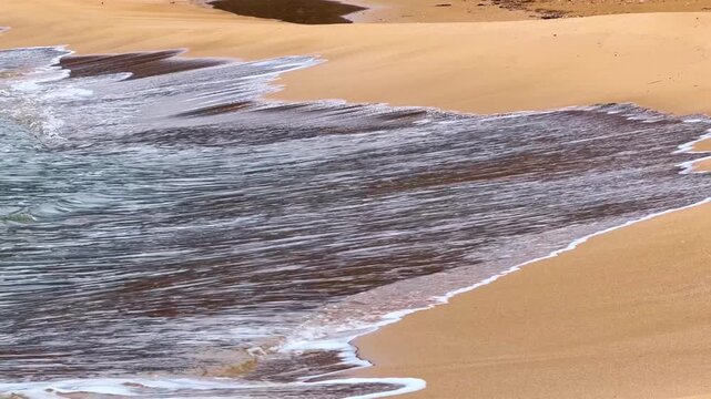 Waves wash onto sandy shore at a beach during daylight hours. Waves roll onto the shore, creating patterns in the sand under sunlight at the beach.