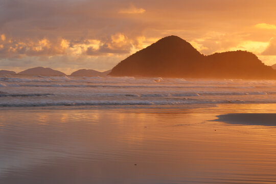 Golden sunrise in Riviera beach, Bertioga, Brazil. Light reflections on sky and water, mountains shilouetttes and cloudy sky. Waves breaking on water edge.