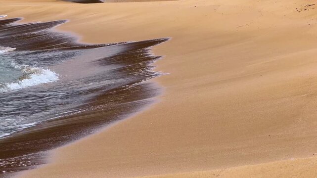 Waves gently touch the sandy shore on a sunny beach in the afternoon. Waves roll onto the shore, leaving patterns in the sand on a warm afternoon at the beach.