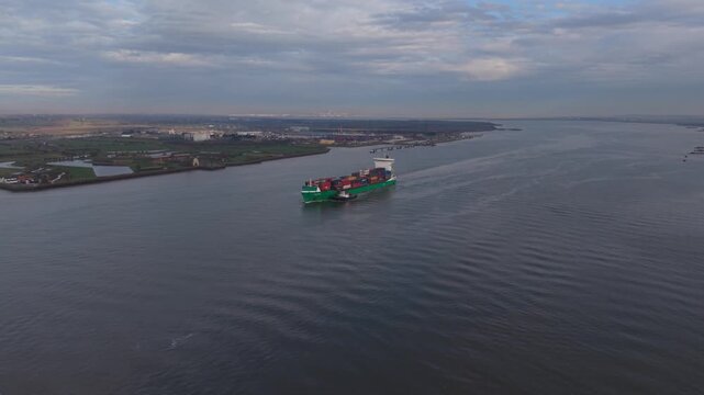 Large container ship sailing on river Thames escorted by pilot boat. High altitude aerial view of industrial landscape with a moving vessel on a cloudy day. 