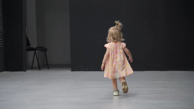 Small child in floral dress begins to crawl then stands and walks away in empty room.