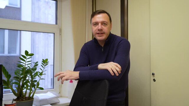 Friendly middle-aged man in dark blue sweater waves hello and then crosses his arms, exuding confidence and approachability in office setting with window and plant.